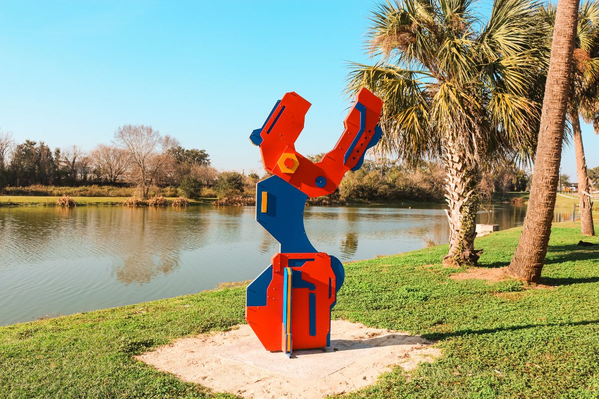 Red and blue abstract sculpture titled “Beckon” by Jeffrey Repko on display near a lake and palm trees at Tuscawilla Park in Ocala.