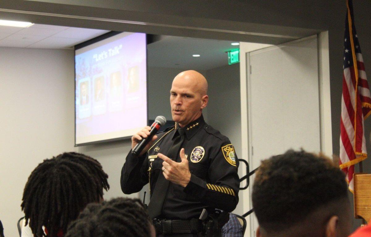 Chief Mike Balken speaking into a microphone at a community forum, with audience members in the foreground.