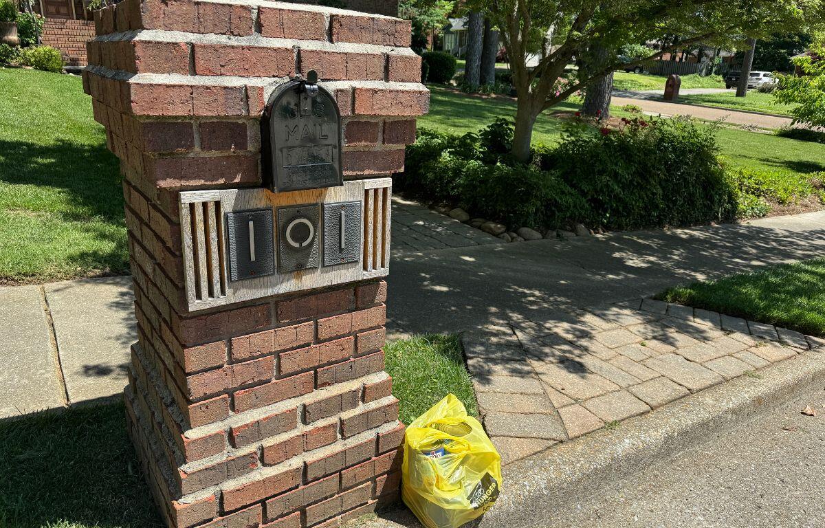 A yellow plastic bag filled with non-perishable food donations sits on the curb next to a brick mailbox structure in a suburban neighborhood, ready for pickup during the Stamp Out Hunger food drive.