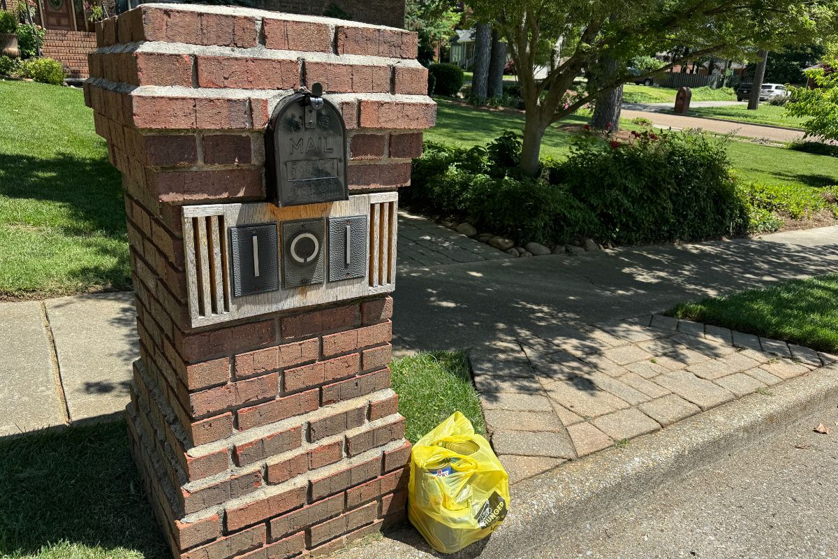 A yellow plastic bag filled with non-perishable food donations sits on the curb next to a brick mailbox structure in a suburban neighborhood, ready for pickup during the Stamp Out Hunger food drive.