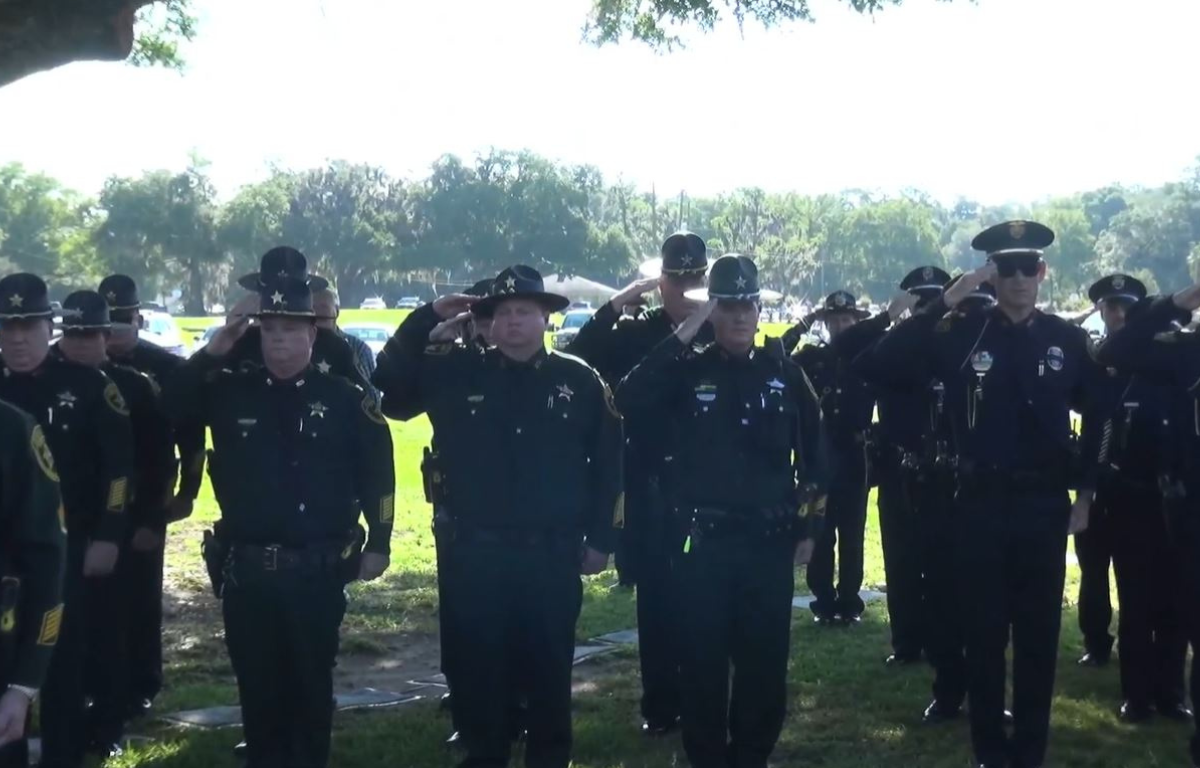 Group of uniformed officers saluting outdoors at a memorial ceremony under a tree.