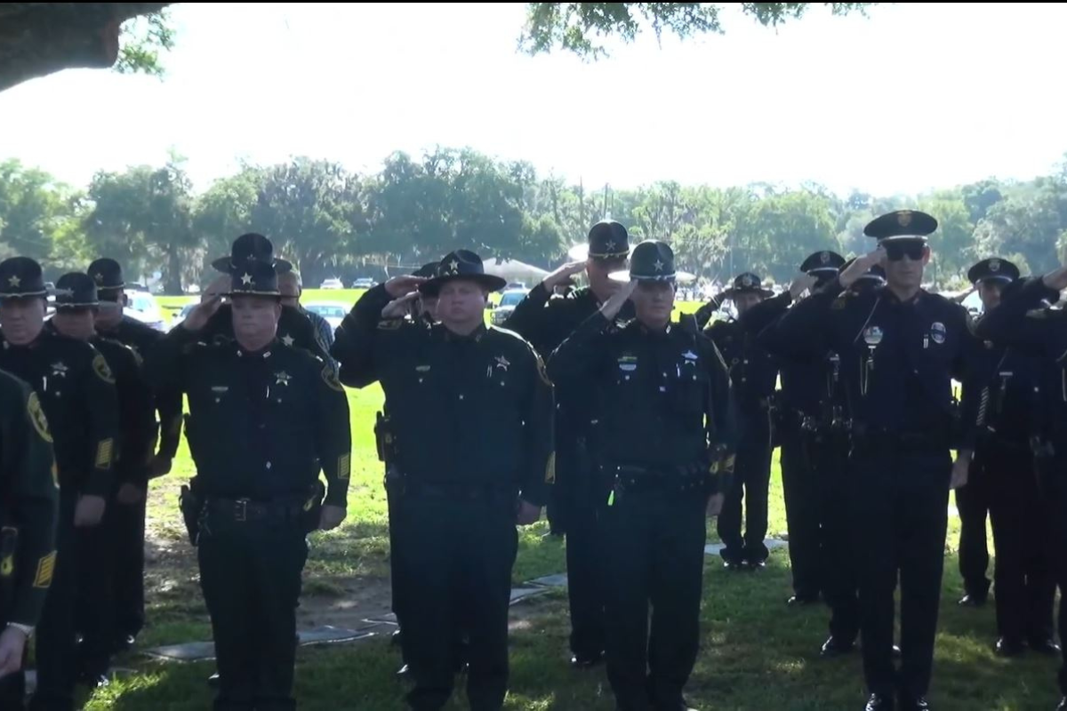 Group of uniformed officers saluting outdoors at a memorial ceremony under a tree.
