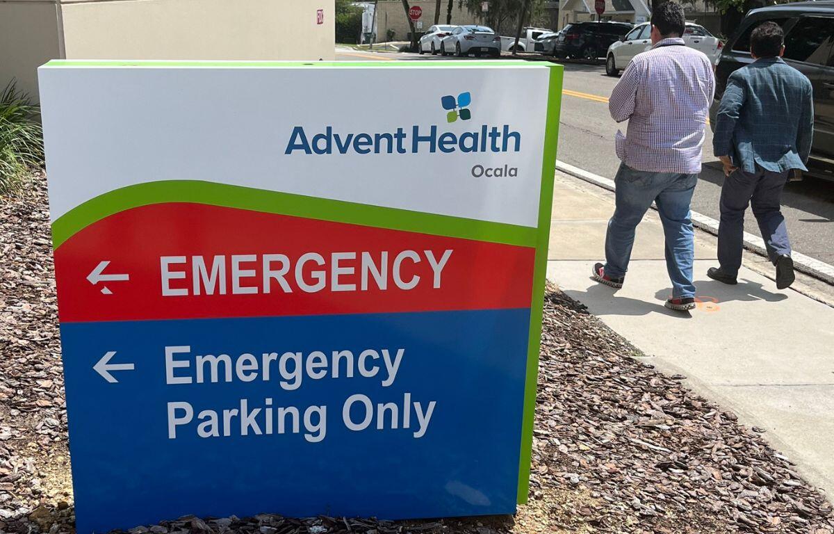 Sign for AdventHealth Ocala emergency department with pedestrians walking nearby on sidewalk.