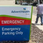 Sign for AdventHealth Ocala emergency department with pedestrians walking nearby on sidewalk.