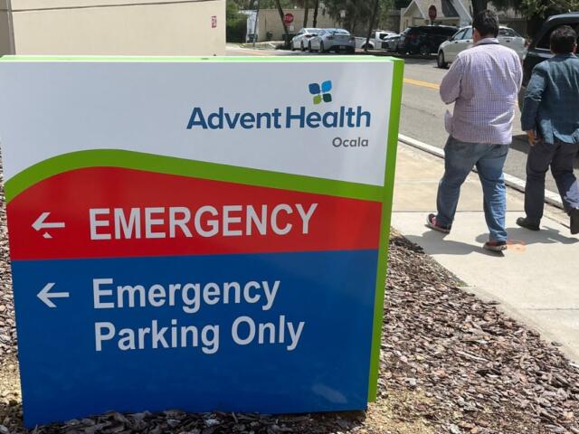 Sign for AdventHealth Ocala emergency department with pedestrians walking nearby on sidewalk.