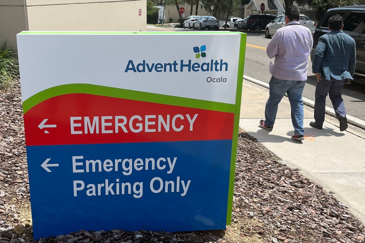 Sign for AdventHealth Ocala emergency department with pedestrians walking nearby on sidewalk.