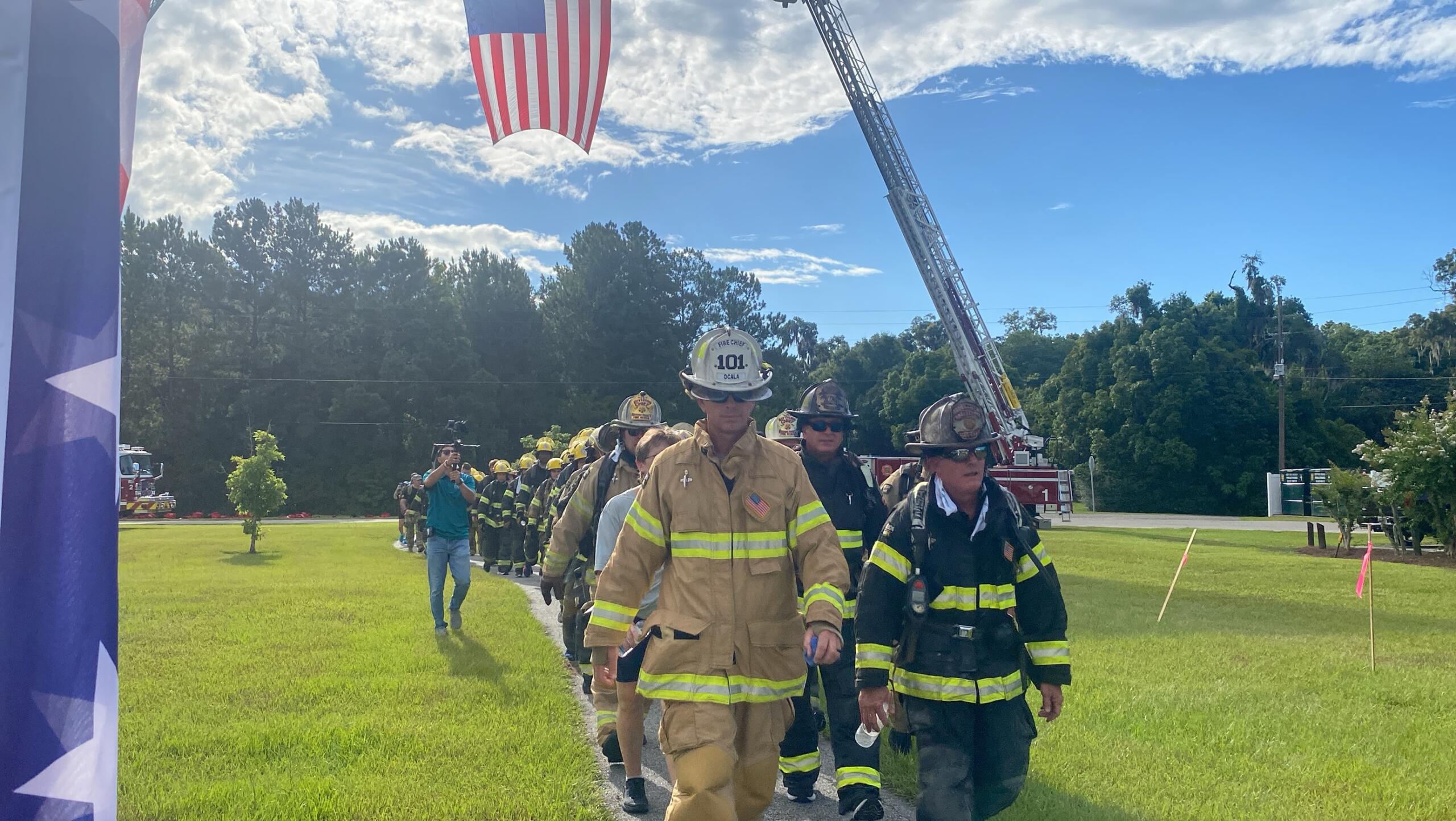 First responders in uniform walk beneath a raised American flag during a PTSD Awareness event at a park in Ocala.