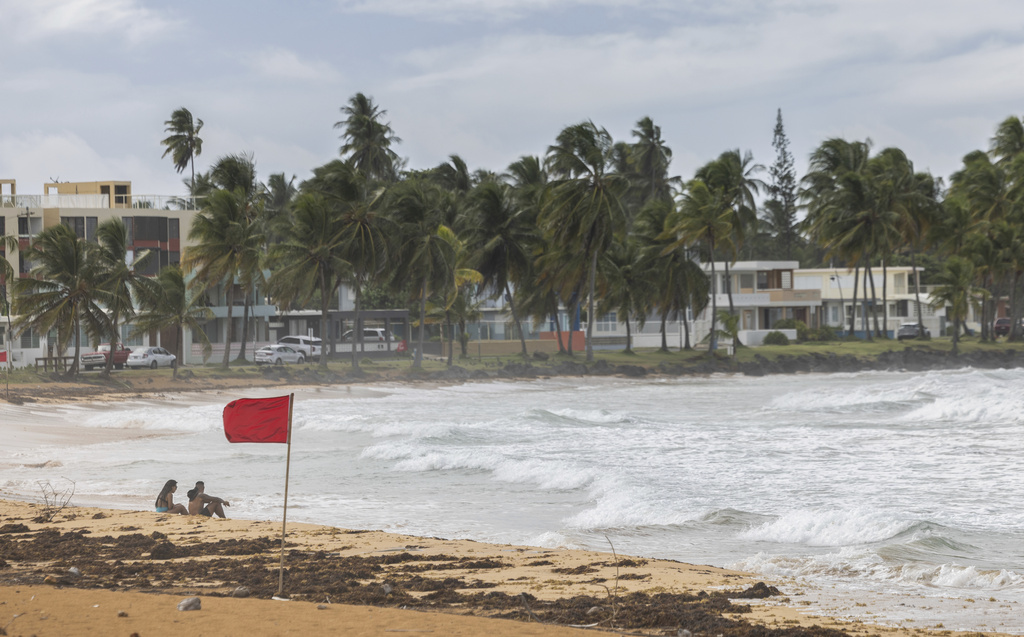 Image shows waves crashing on a beach in Puerto Rico and a hurricane flag standing straight due to high winds.