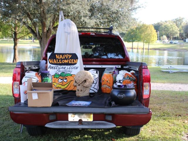 A decorated truck bed featuring Halloween props, including a skull, plastic pumpkins, and a “Happy Halloween” sign from the American Legion Auxiliary, set up beside a lake.