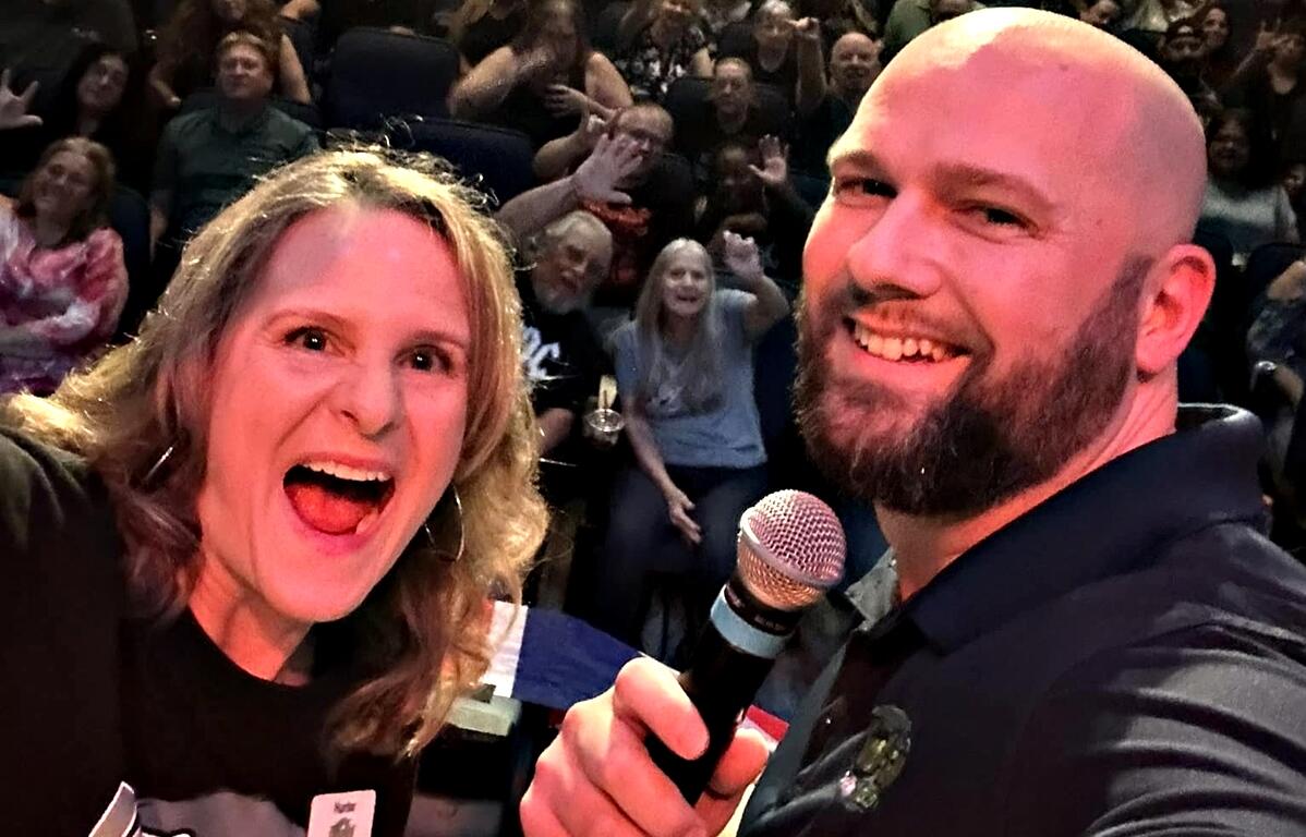 Hunter and Dupree of Wind-FM smiling on stage with a cheering audience behind them at the Reilly Arts Center.