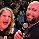 Hunter and Dupree of Wind-FM smiling on stage with a cheering audience behind them at the Reilly Arts Center.