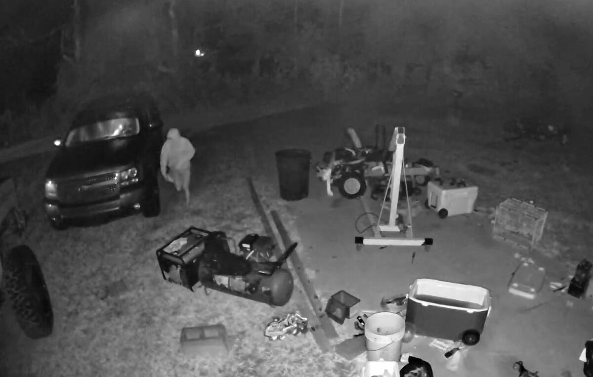 The black and white photo is a still from surveillance video showing a man breaking into a vehicle and taking items from it.