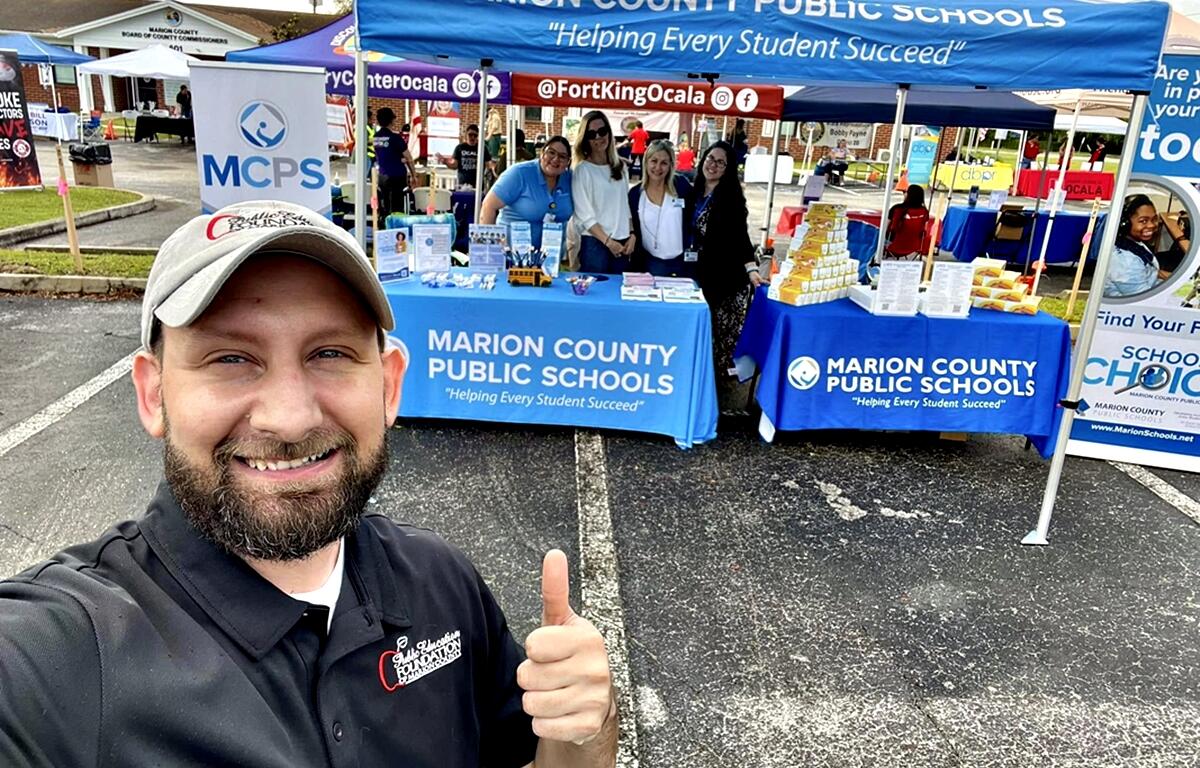 MCPS staff smile under informational tents at a community event with a man in the foreground giving a thumbs up.