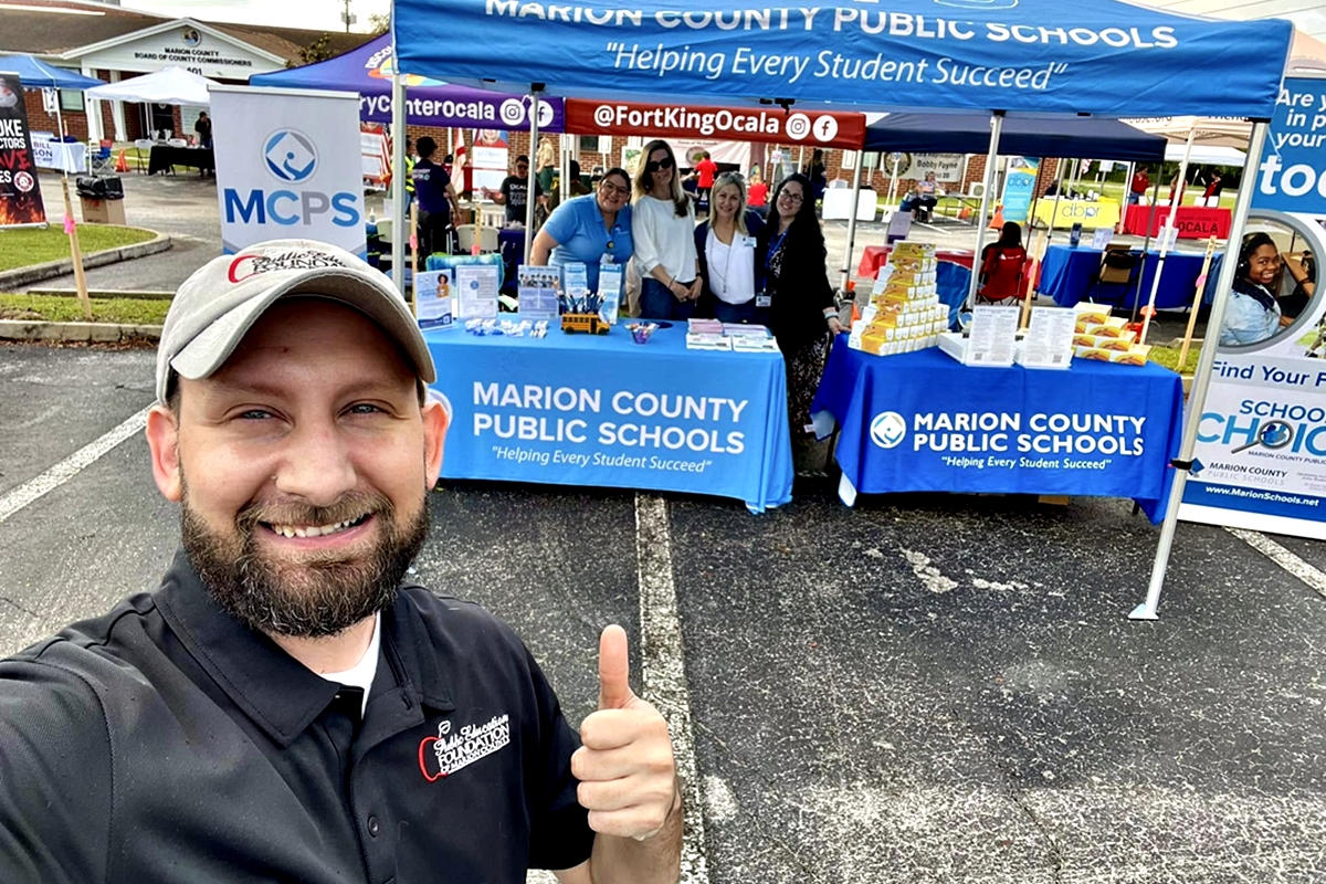 MCPS staff smile under informational tents at a community event with a man in the foreground giving a thumbs up.