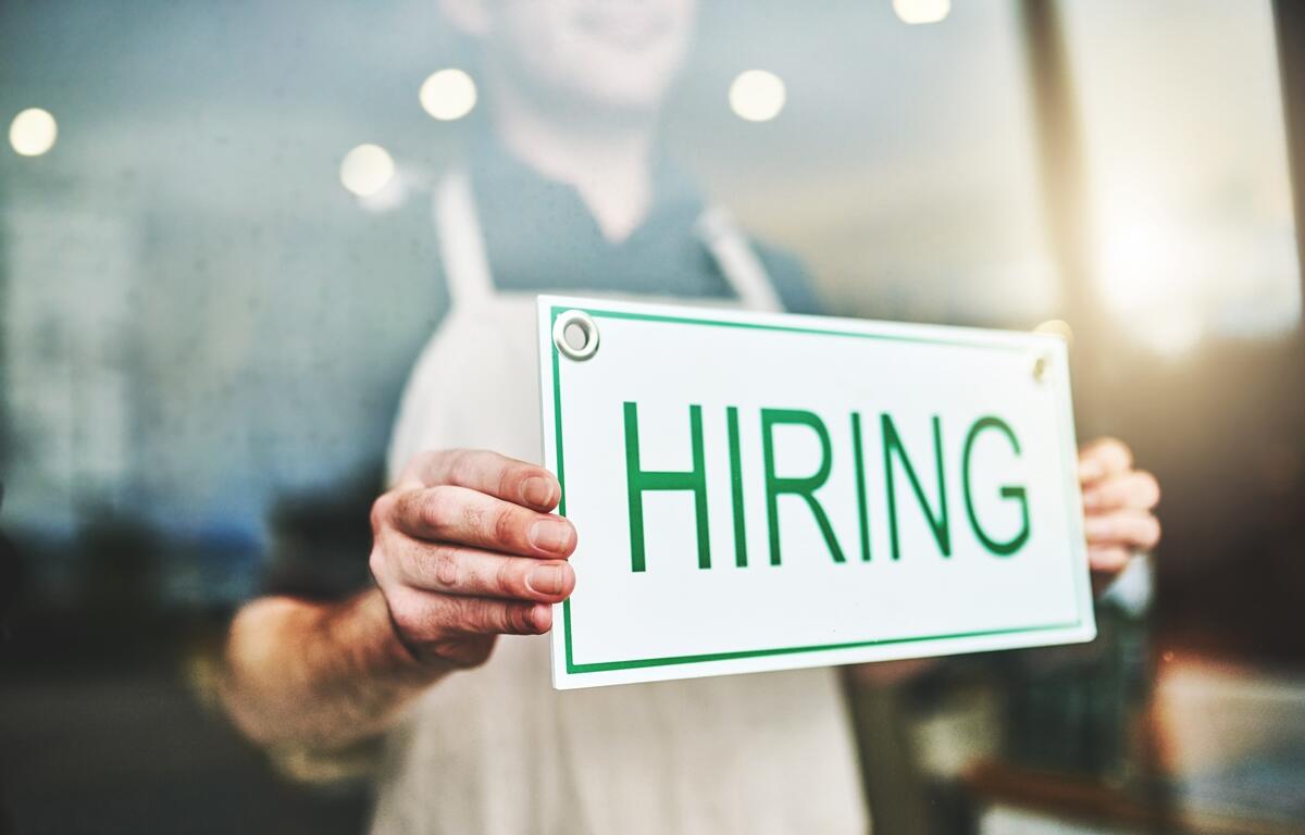 Stock photo of a person holding a “Hiring” sign in a storefront window, symbolizing regional job trends.