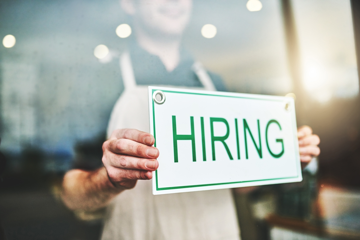 Stock photo of a person holding a “Hiring” sign in a storefront window, symbolizing regional job trends.