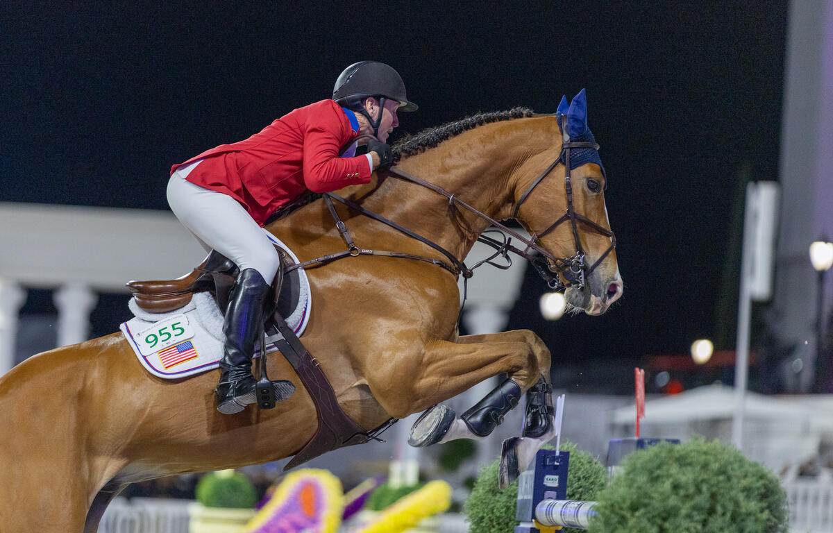 A horse ride wearing ear bonnets, with a rider, in a red blazer, helmet, boots, grasping the reins high up on the withers near the horse's mane, exhibiting a balanced position, with his leg angle and foot position in the stirrups providing the correct cues at the optimal time, as the horses forelegs are poised to go over the vertical, which has flags on top of each standard, with a red flag in the foreground and white flag in the background. And English saddle, sits on top of a saddle, which has a saddle cloth underneath with the riders number and the flag of the United States of America.