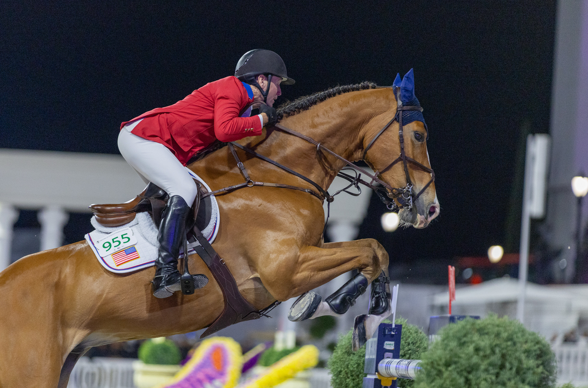 A horse ride wearing ear bonnets, with a rider, in a red blazer, helmet, boots, grasping the reins high up on the withers near the horse's mane, exhibiting a balanced position, with his leg angle and foot position in the stirrups providing the correct cues at the optimal time, as the horses forelegs are poised to go over the vertical, which has flags on top of each standard, with a red flag in the foreground and white flag in the background. And English saddle, sits on top of a saddle, which has a saddle cloth underneath with the riders number and the flag of the United States of America.