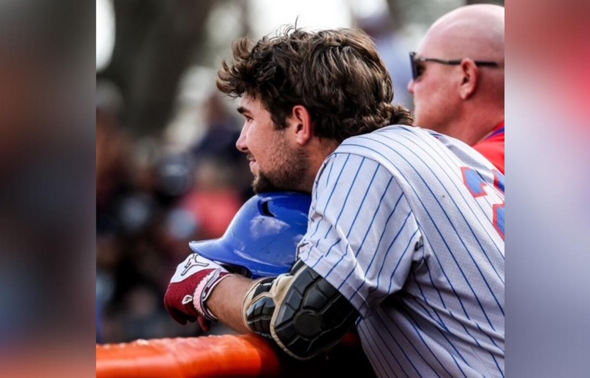 Miles Curley watches from the dugout during a game, resting his helmet on the railing. The CF baseball player contributed to the team's effort in their latest matchup.