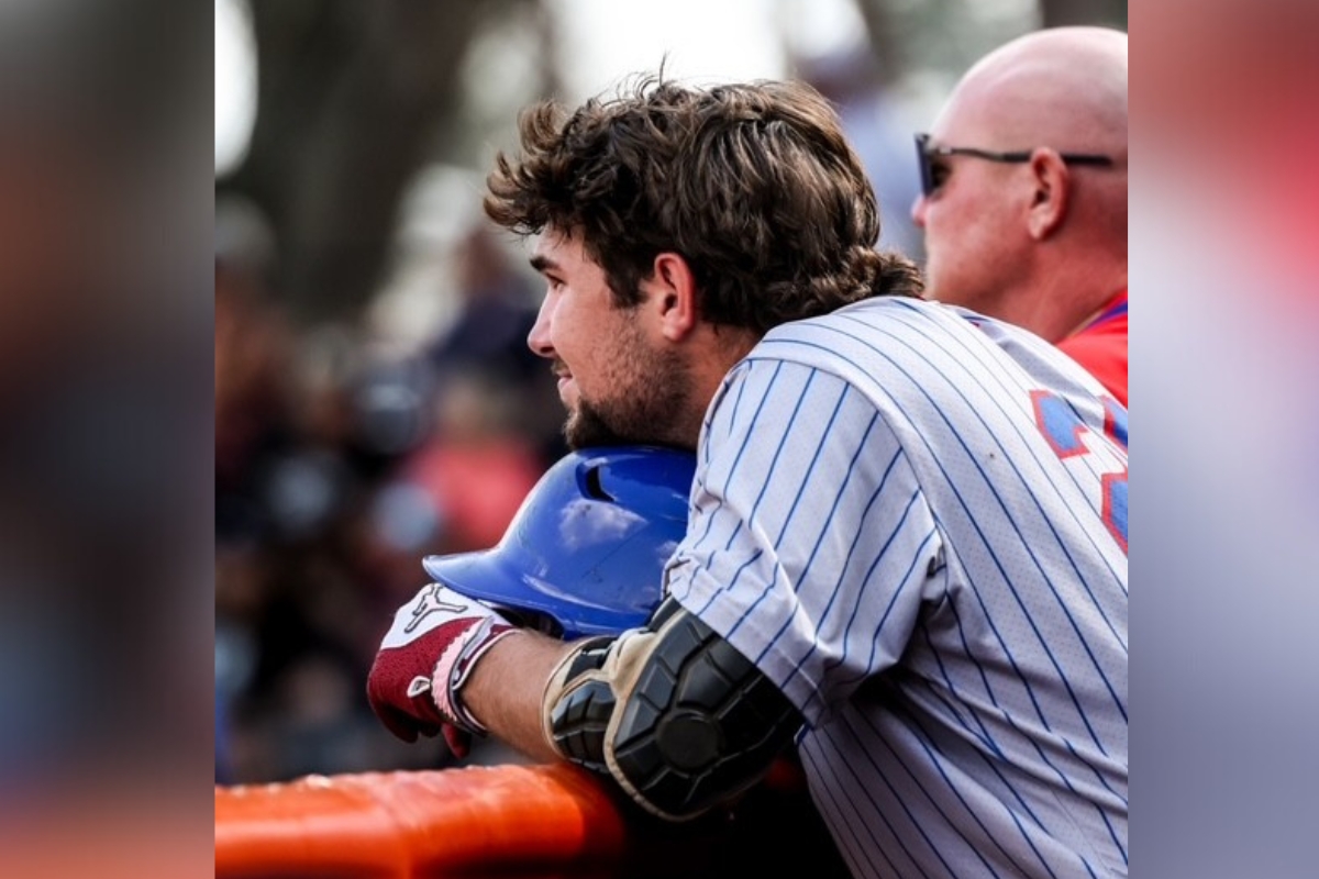 Miles Curley watches from the dugout during a game, resting his helmet on the railing. The CF baseball player contributed to the team's effort in their latest matchup.