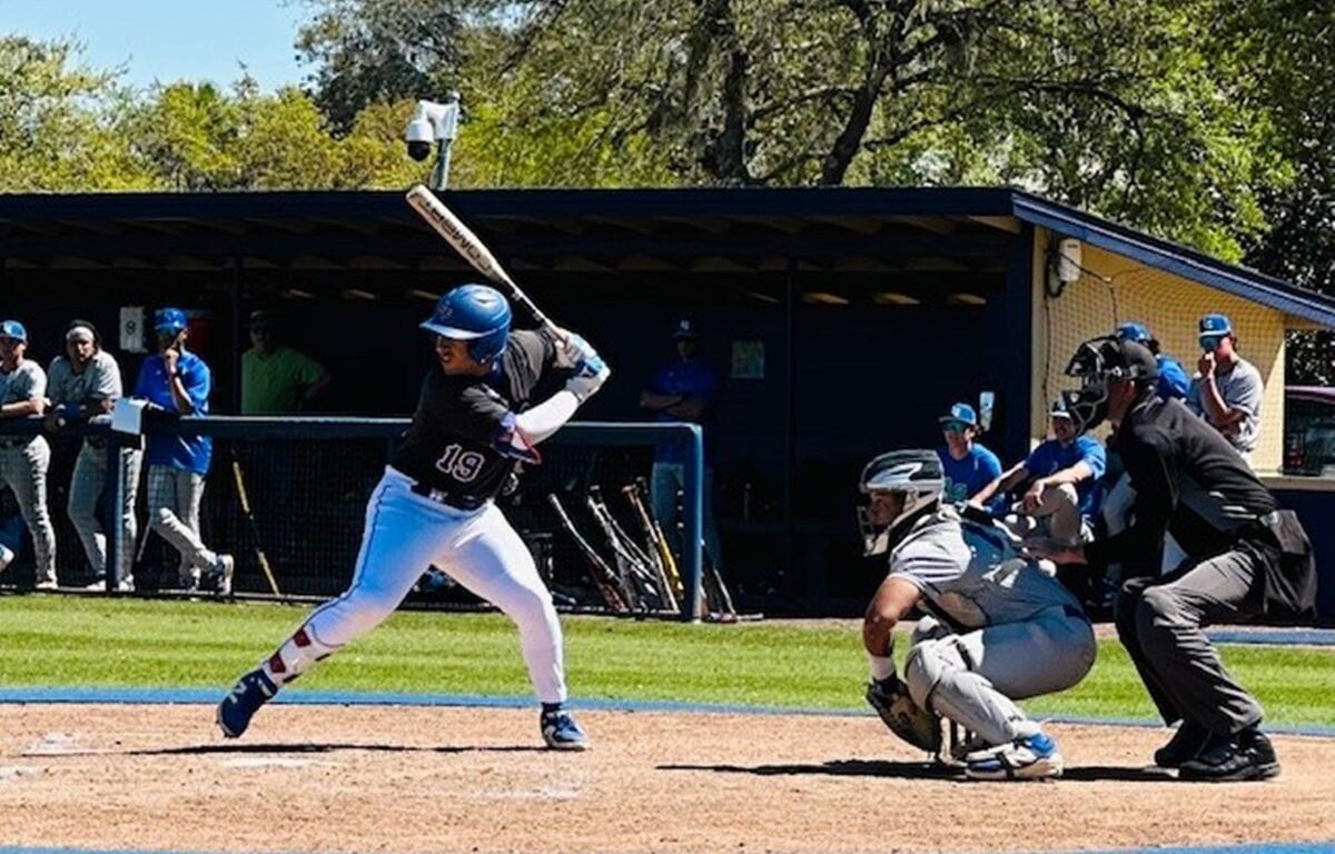 College of Central Florida baseball player Victor Martinez (#19) swings at a pitch during a game as the catcher and umpire watch closely, with teammates and coaches looking on from the dugout in the background.