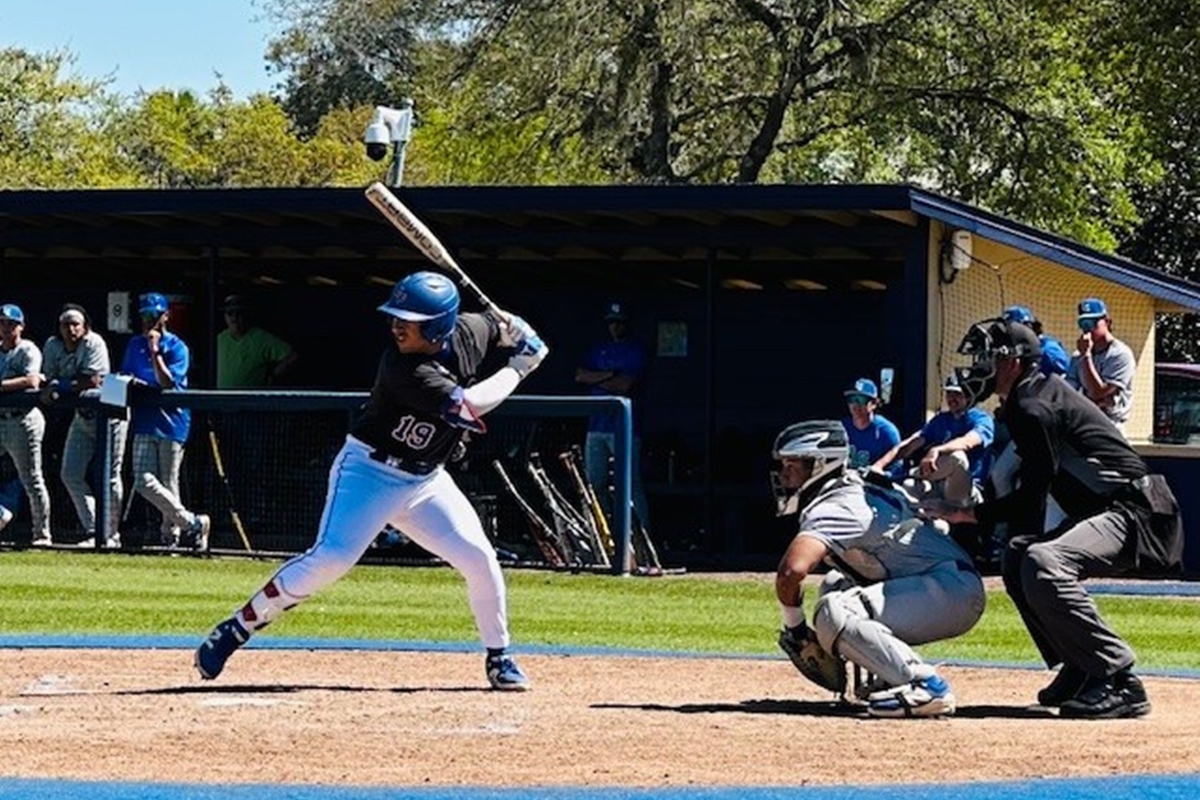 College of Central Florida baseball player Victor Martinez (#19) swings at a pitch during a game as the catcher and umpire watch closely, with teammates and coaches looking on from the dugout in the background.