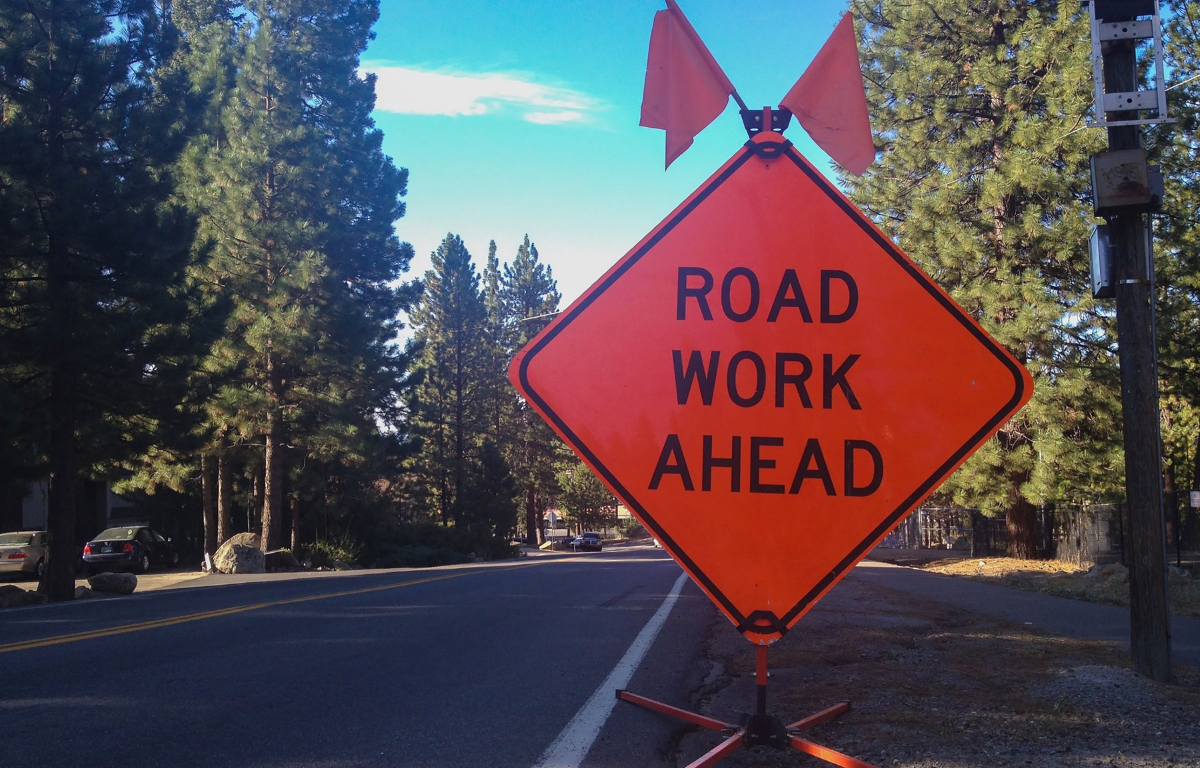 Bright orange "Road Work Ahead" sign with flags on roadside near a tree-lined street.