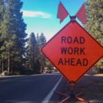 Bright orange "Road Work Ahead" sign with flags on roadside near a tree-lined street.