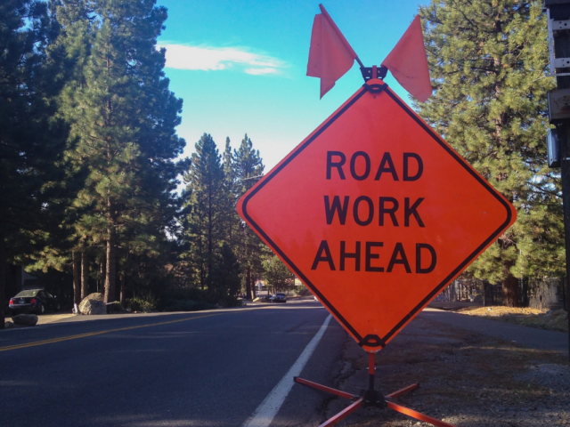 Bright orange "Road Work Ahead" sign with flags on roadside near a tree-lined street.