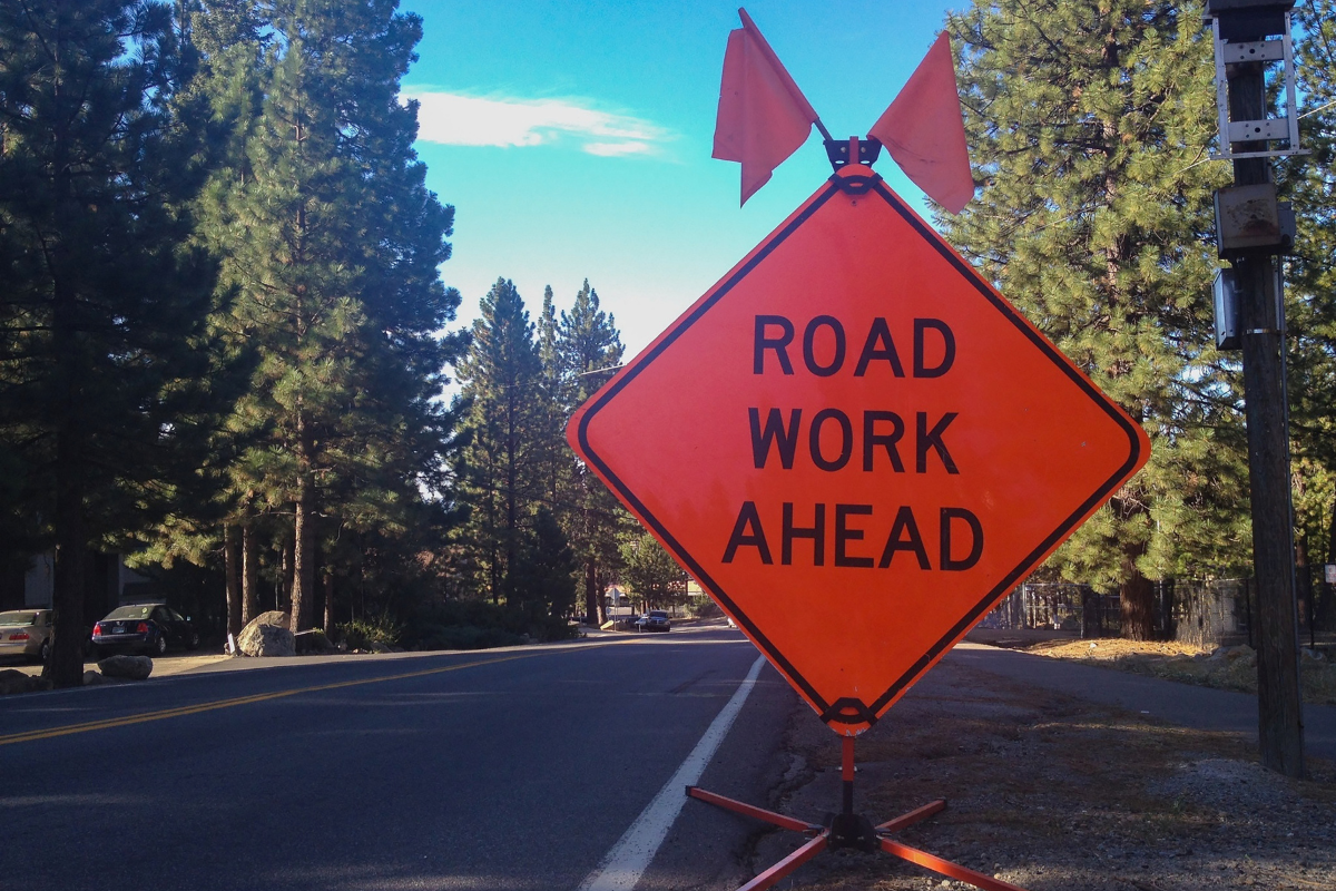 Bright orange "Road Work Ahead" sign with flags on roadside near a tree-lined street.