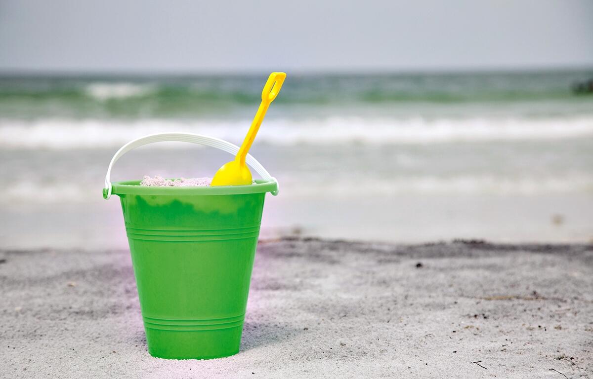 A green bucket and yellow shovel sit on the sand as waves roll in at the beach.