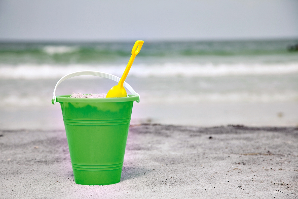 A green bucket and yellow shovel sit on the sand as waves roll in at the beach.