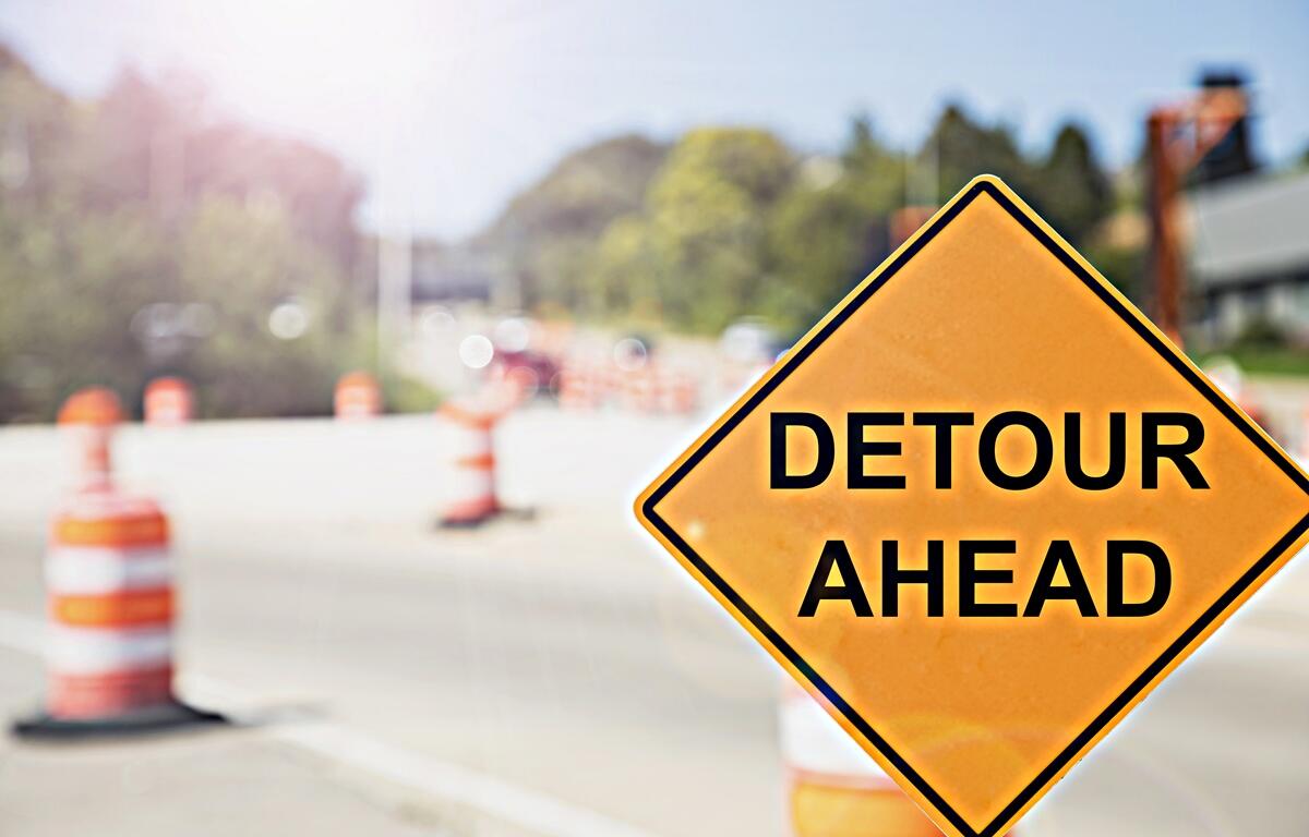 Close-up of a "Detour Ahead" sign with traffic cones and road construction in the background.