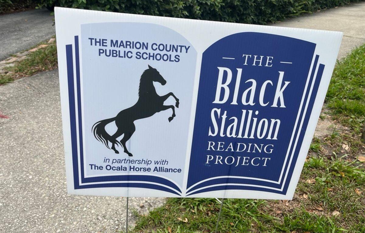 A placard with an image of a horse and writing in the left, and writing against a blue background on right. On the left is pavement in the background and foreground, and on the right is grass.
