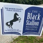 A placard with an image of a horse and writing in the left, and writing against a blue background on right. On the left is pavement in the background and foreground, and on the right is grass.