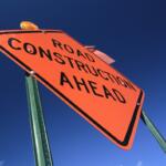 Close-up of an orange “Road Construction Ahead” warning sign against a clear blue sky.
