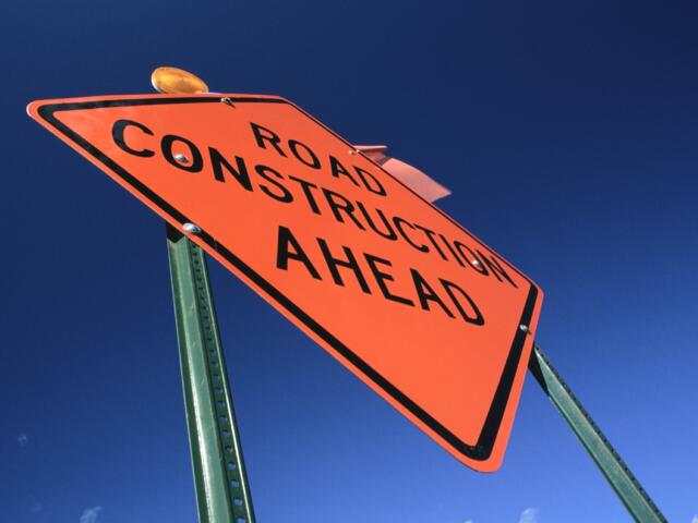Close-up of an orange “Road Construction Ahead” warning sign against a clear blue sky.