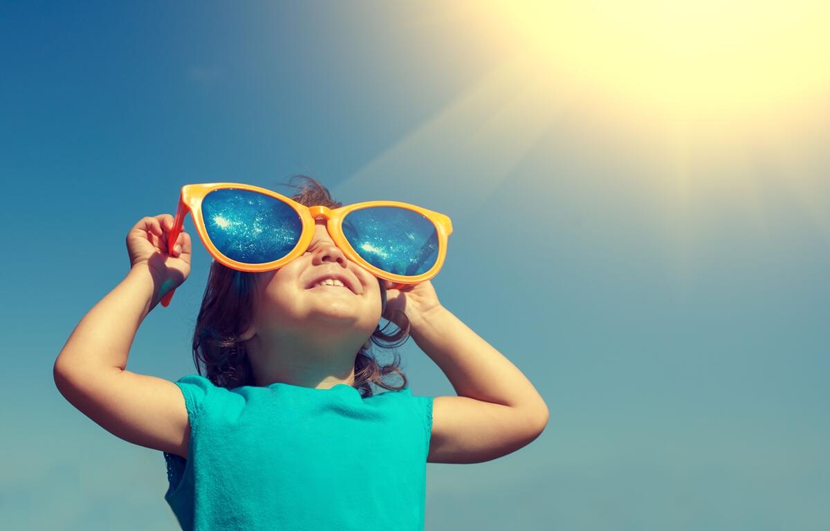 A child wearing oversized sunglasses smiles under the sun on a clear, warm day.