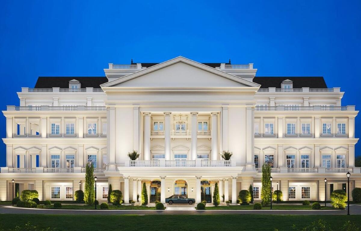 A grand, illuminated building with classical architecture, featuring white columns and multiple windows, set against a clear blue evening sky, with a luxury car parked in front and well-maintained greenery.
