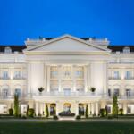 A grand, illuminated building with classical architecture, featuring white columns and multiple windows, set against a clear blue evening sky, with a luxury car parked in front and well-maintained greenery.