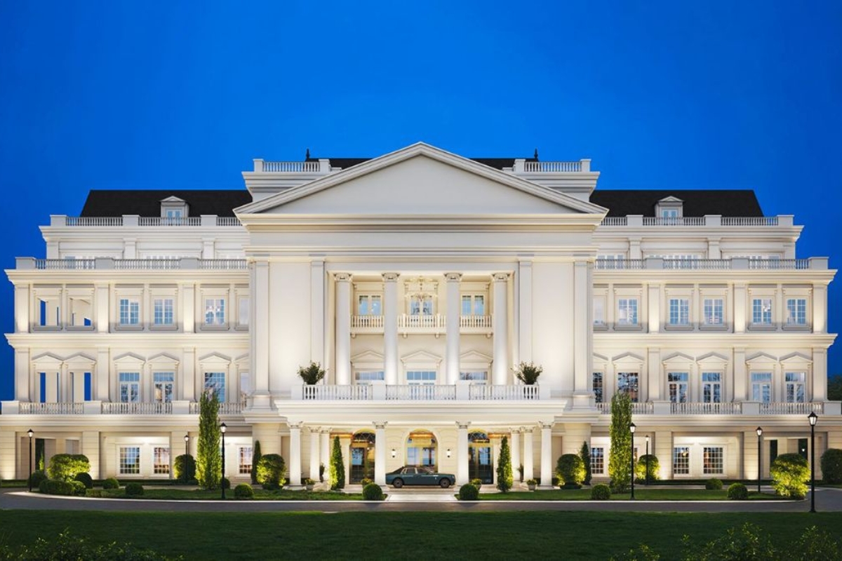 A grand, illuminated building with classical architecture, featuring white columns and multiple windows, set against a clear blue evening sky, with a luxury car parked in front and well-maintained greenery.