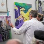 A man in a white shirt gestures toward a colorful painting of a saxophone player on a gallery wall while speaking with a woman. Around them, several vibrant artworks are displayed, including a painting of a violinist under a tree and abstract pieces with musical themes. A blue ribbon labeled "1st Place" hangs beside one of the paintings, indicating award recognition. Other attendees are partially visible in the foreground.
