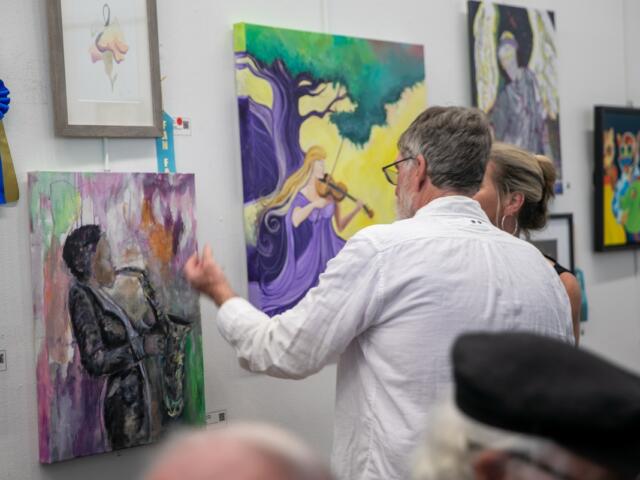 A man in a white shirt gestures toward a colorful painting of a saxophone player on a gallery wall while speaking with a woman. Around them, several vibrant artworks are displayed, including a painting of a violinist under a tree and abstract pieces with musical themes. A blue ribbon labeled "1st Place" hangs beside one of the paintings, indicating award recognition. Other attendees are partially visible in the foreground.