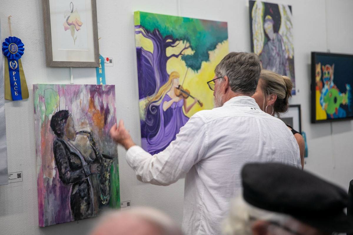 A man in a white shirt gestures toward a colorful painting of a saxophone player on a gallery wall while speaking with a woman. Around them, several vibrant artworks are displayed, including a painting of a violinist under a tree and abstract pieces with musical themes. A blue ribbon labeled "1st Place" hangs beside one of the paintings, indicating award recognition. Other attendees are partially visible in the foreground.