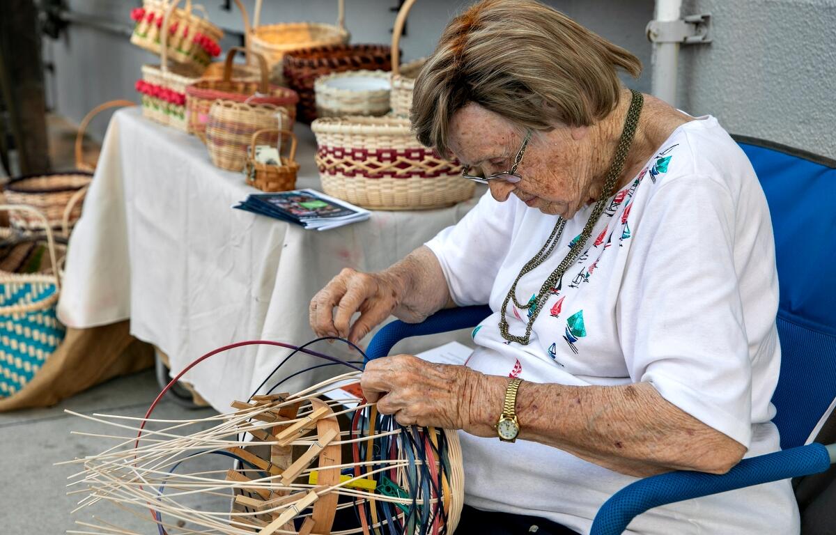 An elderly woman with light hair and glasses sits in a blue chair, carefully weaving a colorful basket by hand. She is surrounded by a display table filled with completed woven baskets in various patterns and sizes.