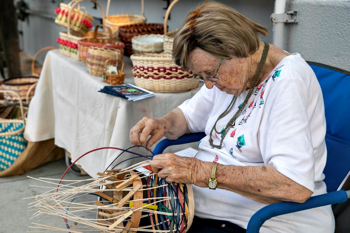 An elderly woman with light hair and glasses sits in a blue chair, carefully weaving a colorful basket by hand. She is surrounded by a display table filled with completed woven baskets in various patterns and sizes.