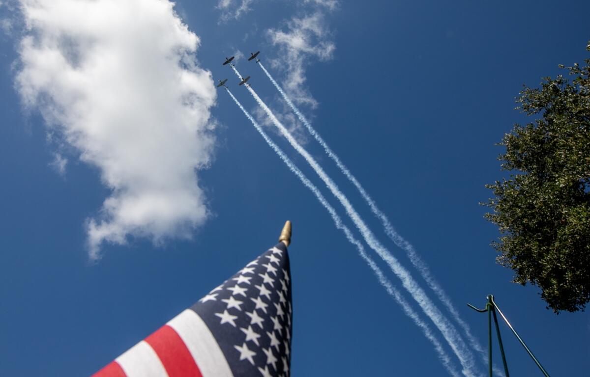 Three planes leave smoke trails in a Memorial Day flyover above an American flag and tree branches against a blue sky.