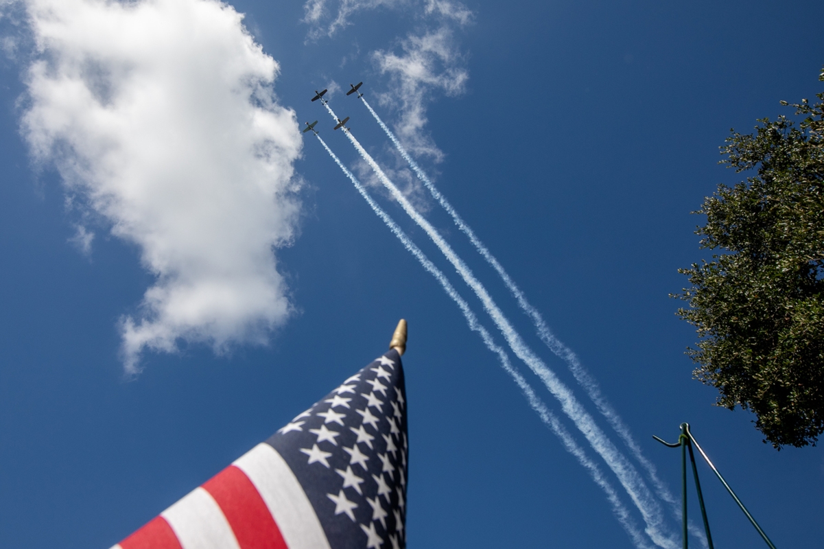 Three planes leave smoke trails in a Memorial Day flyover above an American flag and tree branches against a blue sky.