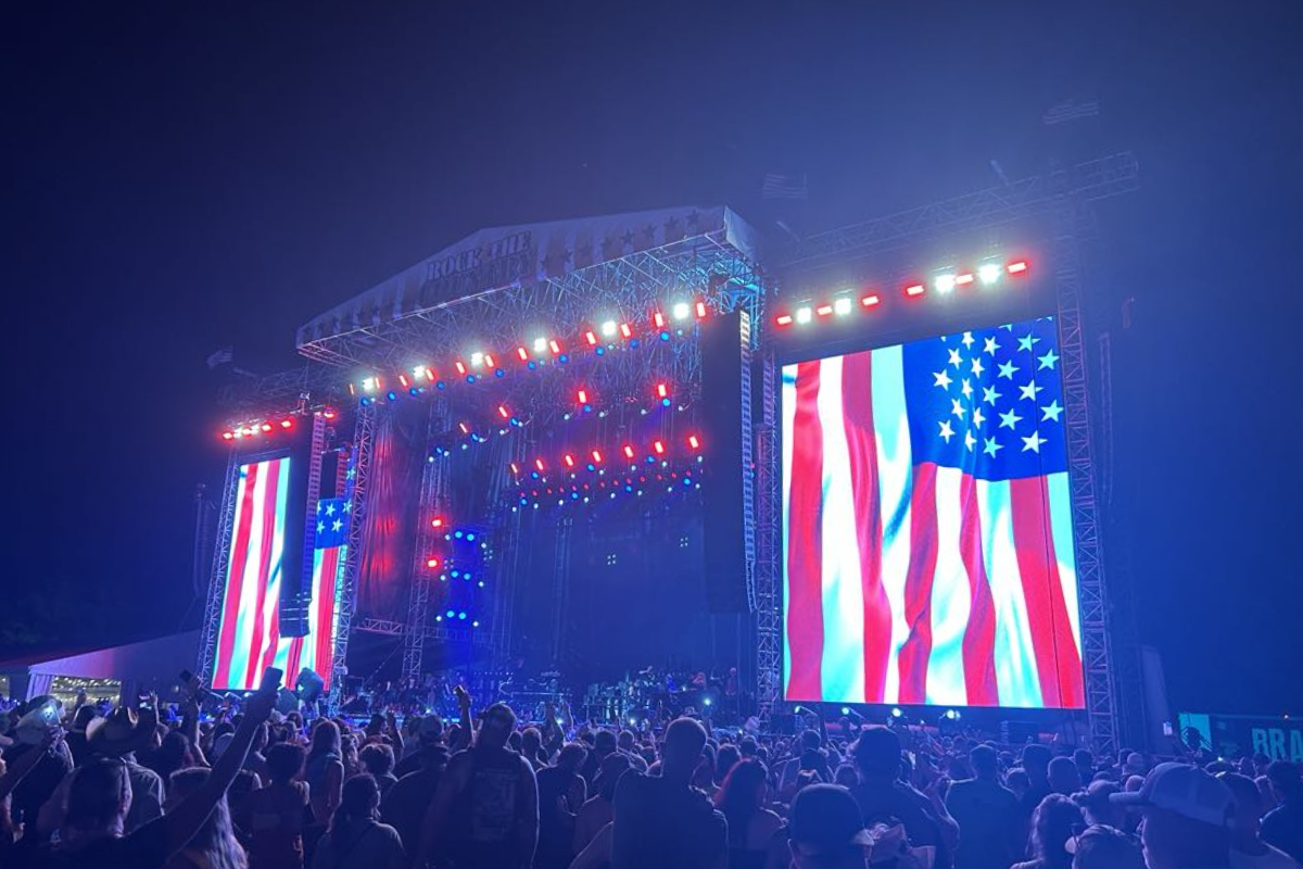 A large nighttime concert crowd faces a brightly lit stage displaying American flags on two giant vertical screens, with red, white, and blue lights illuminating the scene.