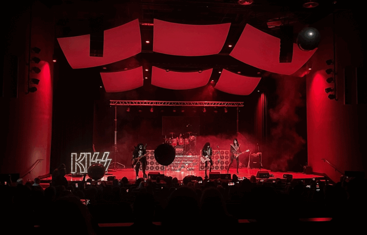 A KISS tribute band performs on stage under dramatic red lighting, with smoke effects and large black balloons floating above the audience. The band members wear classic KISS makeup and costumes, and the stage is decorated with illuminated 'KISS' lettering and stacked speaker props.